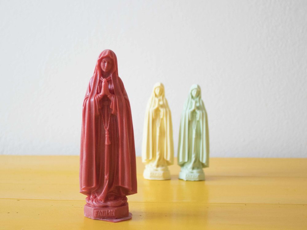Três imagens de Nossa Senhora de Fátima, onde a que está destacada está mais à frente e é vermelha.